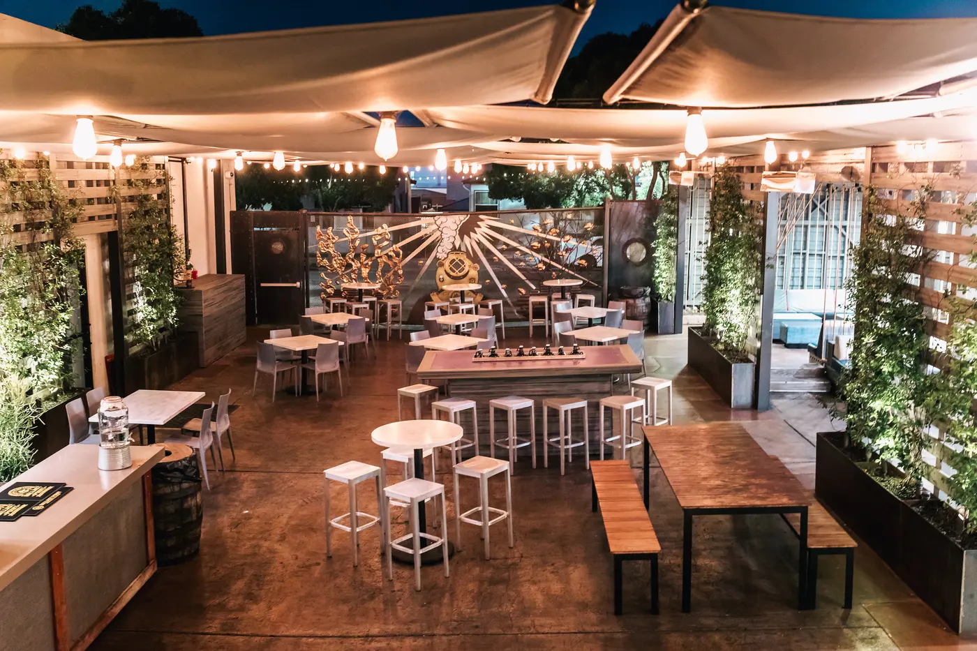 The Ballast Point Little Italy patio at dusk: string lights, bar tables and stools under canvas shades